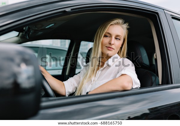 Woman driver with\
long blond hair dressed in white shirt and black skirt on the\
parking is sitting in the car. She is looking at camera with great\
emotions. Sunny day. Work\
time.