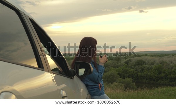 Woman driver traveler resting, drinking morning\
coffee from a mug in the sun. A tourist girl holds mug of hot tea\
in her hands and looks at sunset next to car. Freedom of travel and\
tourism by car.
