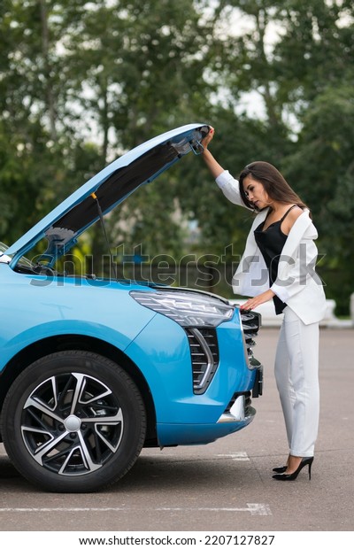 Woman driver tries to figure out\
problem looking under blue car open hood. Successful lady wants to\
repair car alone without car services on parking\
lot