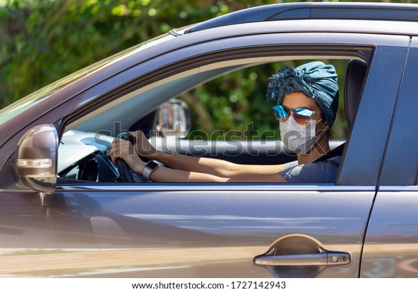 A woman driving a car wearing a mask, scarf in her hair\
and sunglasses to protect herself from contamination by corona\
virus. Pandemic. Essential services. Social isolation. Health care.\

