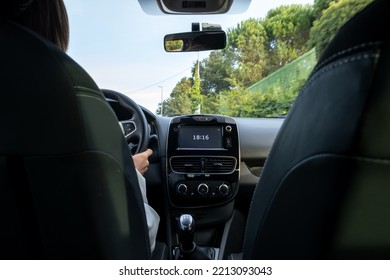 Woman Driving, View From The Back Seat At A Low Angle To The Car Main Dashboard With Screen Clock With 18:16 Time Showing. From The Back Rear Passenger Seat POV.