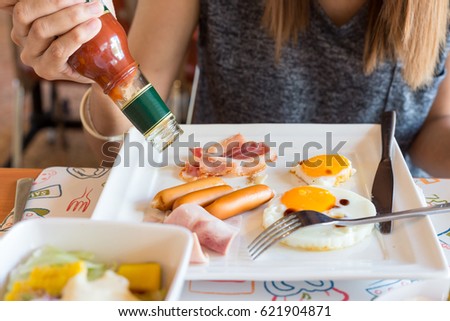 woman eat breakfast, she pouring tomato sauce or ketchup sauce bottle on sausage, ham and fried egg