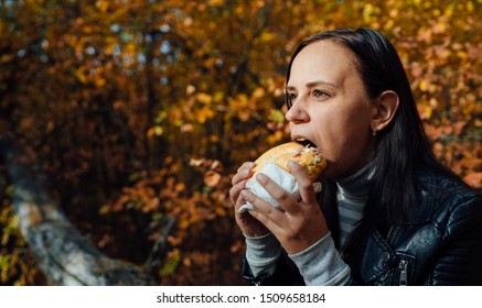 Woman eating a Burger in a sunny spring park. Young woman with Sunny day eating hamburger in autumn forest.