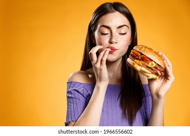 Woman eating cheeseburger with satisfaction. Girl enjoys tasty hamburger takeaway, licking fingers delicious bite of burger, order fastfood delivery while hungry, standing over orange background