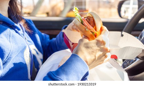 A Woman Is Eating Chicken Sandwich Which She Got From A Roadside Drive Through Fast Food Chain. She Has Packs Of Food On Her Lap As She Eats In The Driver Seat Of Her Car In The Parking Lot.