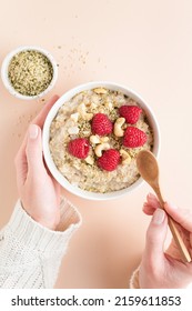 Woman Eating Cooked Oatmeal Porridge With Hemp Seeds, Cashew Nuts And Fresh Raspberries. Top View. Female Hands Holding Healthy Breakfast Oatmeal Bowl