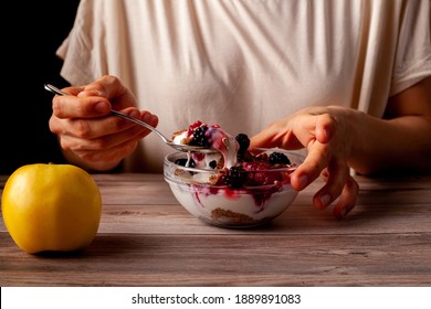 Woman Is Eating A Fresh Home Made Glass Bowl Of Creamy Yogurt Parfait With Berries, Muesli And Seeds As Late Night Snack. Photo Shows Hand Holding A Spoon With An Apple On Side. Background Is Dark.