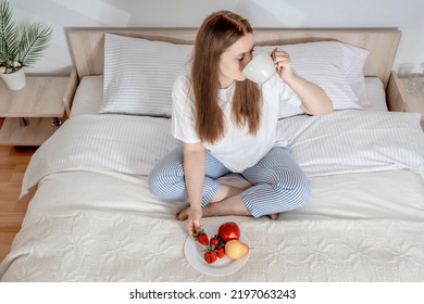 Woman eating fruit breakfast in bed at home in the morning. Lady with long hair in pajamas. Girl on clean white bedding with cozy blanket. Healthy lifestyle, balance, wellness, time for yourself. - Powered by Shutterstock