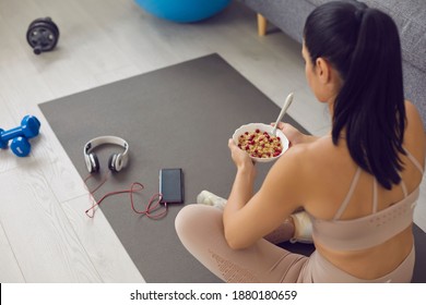 Woman Eating Healthy Food After Fitness Workout At Home. Fit Young Female Sitting On Sports Mat In The Living-room, Relaxing And Having Bowl Of Natural Cranberry Granola. High Angle Over Shoulder View