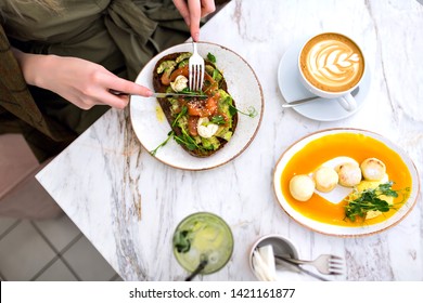 Woman Eating Her Tasty Brunch On Hipster Cafe, Top View Of Marble Table, Salmon Avocado Toast , Coffee And Sweet Tasty Cheesecakes, Enjoying Her Breakfast.