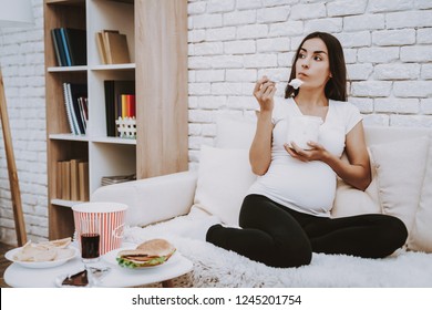 Woman Is Eating A Ice Cream With Spoon. Woman Is A Young Brunette Pregnant Girl. Girl Sitting On Couch. Woman Is Thinking. Different Food And Cola On Table. Person Located At Home.