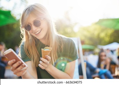 Woman eating icecream using smartphone. Portrait of a girl with ice cream browsing through social media or messaging her friends enjoying summer in the city park. - Powered by Shutterstock