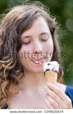 Woman eating one vanilla chocolate ice cream gelato cone looking down tongue licking with bokeh background of park in Europe city during sunny summer day