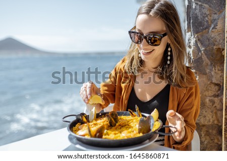 Woman eating paella, traditional spanish dish, while sitting at the restaurant terrace near the ocean. Concept of sea food and good summer vacations