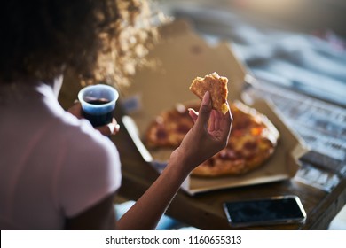 woman eating pizza and drinking cola while sitting on sofa watching tv in home late at night