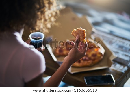 woman eating pizza and drinking cola while sitting on sofa watching tv in home late at night