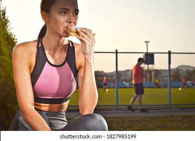 Woman Eating A Sandwich After A Workout. Athlete Resting On A Background Of Sunset.