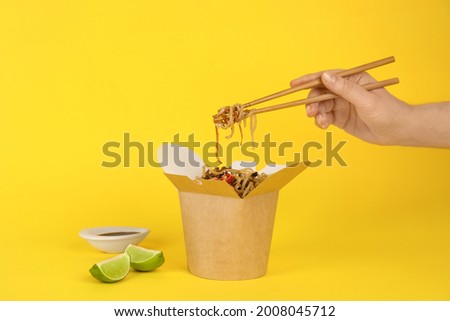 Woman eating seafood wok noodles with chopsticks from box on yellow background, closeup