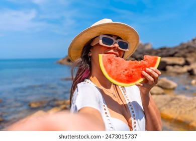 A woman is eating a watermelon on a beach. The watermelon is cut in half and she is holding it in her hand. The beach is a beautiful and relaxing setting, with the ocean in the background - Powered by Shutterstock