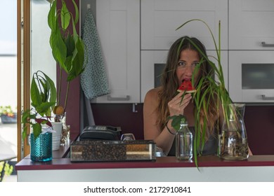 woman eats watermelon in the kitchen behind the counter. In front of the woman on the counter are flowers and coffee beans in a glass jar. Behind the woman are white kitchen cabinets.