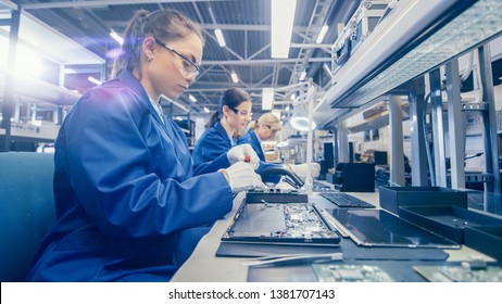 Woman Electronics Factory Worker In Blue Work Coat And Protective Glasses Is Assembling Laptop's Motherboard With A Screwdriver. High Tech Factory Facility With Multiple Employees.