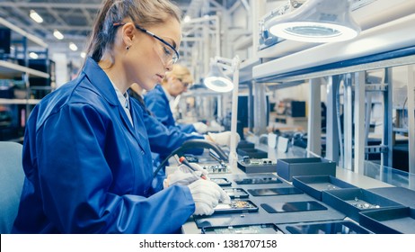 Woman Electronics Factory Worker in Blue Work Coat and Protective Glasses is Assembling Smartphones with Screwdriver. High Tech Factory Facility with more Employees in the Background. - Shutterstock ID 1381707158
