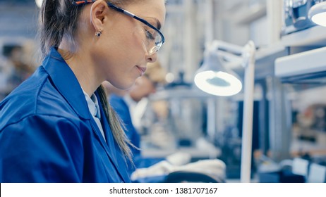 Woman Electronics Factory Worker In Blue Work Coat And Protective Glasses Is Assembling Smartphones With Tweezers And Screwdriver. High Tech Factory Facility With More Employees In The Background.