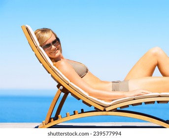 Woman In Elegant Bikini Relaxing Beside A Swimming Pool On Wood Wicker Chair