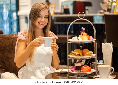 Woman in elegant dress enjoy English breakfast Afternoon tea in modern restaurant. Girl eating delicious sweet food. Cakes, sandwiches and macarons on high tea stand.