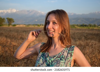 Woman in elegant dress on field full of ripe rye holds a rye stem in her hand and mouth. Summer portrait of redhead or brunette female on a wheat field at sunset, harvest time. Pretty woman portrait. 