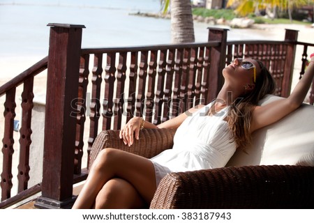 Woman in elegant dress relaxing at resort terrace enjoying luxury lifestyle outdoor day dreaming. Female relaxing on lounge chair. Vacations And Tourism Concept.