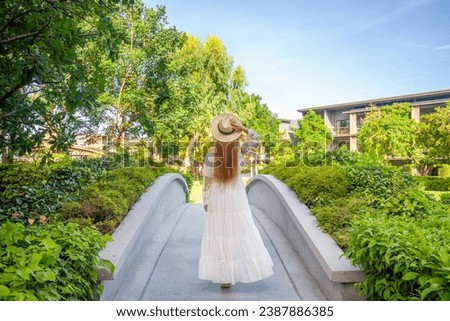 Woman in elegant white dress strolls through modern Thai hotel, immersed in lush greenery. Thailand vacation bliss.