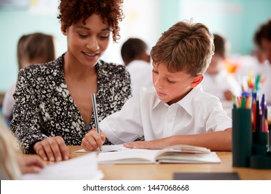 Woman Elementary School Teacher Giving Male Pupil Wearing Uniform One To One Support In Classroom