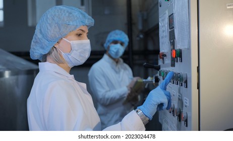 A Woman, An Employee Of An Enterprise Or A Factory, In A Sterile White Coat, A Medical Mask, A Disposable Cap, Hands In Disposable Latex Gloves, Presses The Equipment Button, A Colleague In The