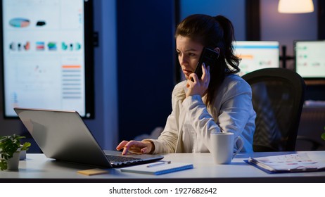 Woman employee speaking at phone while working at laptop late at night. Busy focused freelancer using modern technology network wireless doing overtime for job reading writing, searching taking break - Shutterstock ID 1927682642