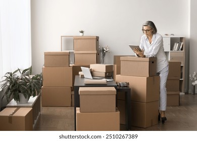 Woman engaged in managing orders and inventory for e-commerce or small business, working in stockroom using tablet, check, update order information. Labeling, packaging, dispatching parcels workflow - Powered by Shutterstock