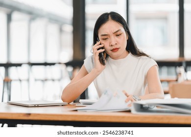 woman is engaged in phone call while reviewing documents at desk. She appears focused and slightly concerned, surrounded by modern workspace. - Powered by Shutterstock