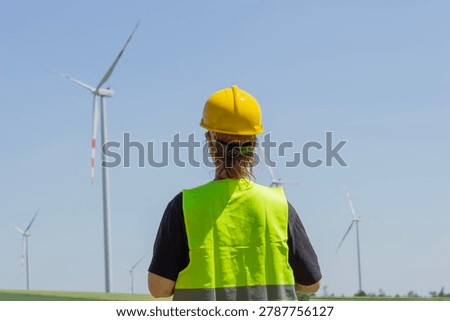 woman engineer back view wind turbine looking away yellow hard hat protective vest inspection green field sunny atmosphere concept of renewable energy, clean power, sustainable industry