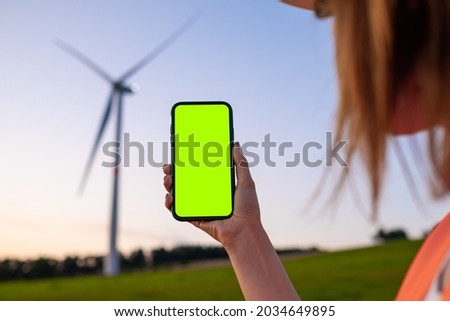 Woman engineer holding a smartphone with chromo key on the background of wind turbine at sunset.