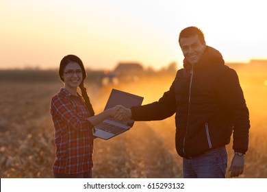 Woman engineer with laptop and landowner shaking hands on field at sunset with tractor working in background. Agribusiness concept