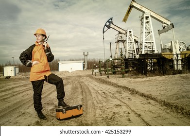 Woman engineer in the oil field talking on the radio wearing orange helmet and work clothes. Industrial site background. Toned.