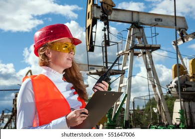 Woman engineer in the oil field talking on the radio wearing red helmet and orange work clothes. Industrial site background. Oil and gas concept. 