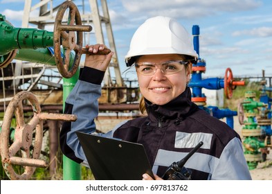 Woman Engineer In The Oilfield Near Wellhead Wearing White Helmet And Work Clothes. Industrial Site Background. Oil And Gas Concept.