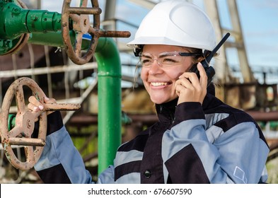 Woman Engineer In The Oilfield Talking On The Radio Wearing White Helmet And Work Clothes. Industrial Site Background. Oil And Gas Concept.