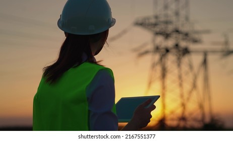 Woman Engineer, Power Engineer In Helmet Checks Power Line Using Computer Tablet Online. Electrician In Outdoors. Electric Lines Of High Voltage At Sunset. Distribution And Supply Of Electricity