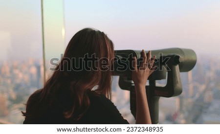 Woman enjoy Bangkok cityscape on Baiyoke Tower Sky viewpoint. Girl looking in binoculars telescope on observation deck for tourist. City skyline panorama at sunset. Travel, tourism, visit Thailand