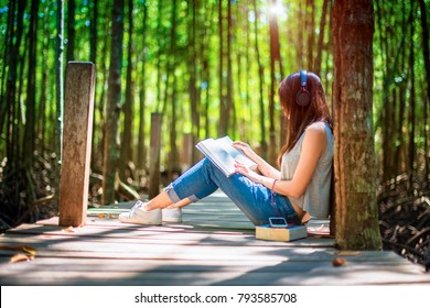 Woman Enjoy Reading On Wooden Bridge Under Shadow And Light Of Forest Tree, Feeling Relax And Comportatble Easy Listening Music Online From Mobile Device In The Meanwhile