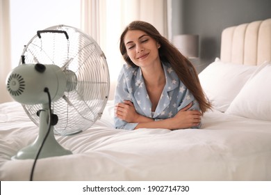 Woman enjoying air flow from fan on bed in room. Summer heat
