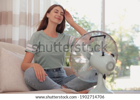 Woman enjoying air flow from fan on sofa in living room. Summer heat