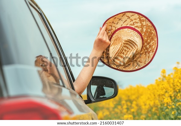 Woman enjoying car ride in blooming summer\
countryside landscape, hand with straw hat reaching out the window,\
selective focus